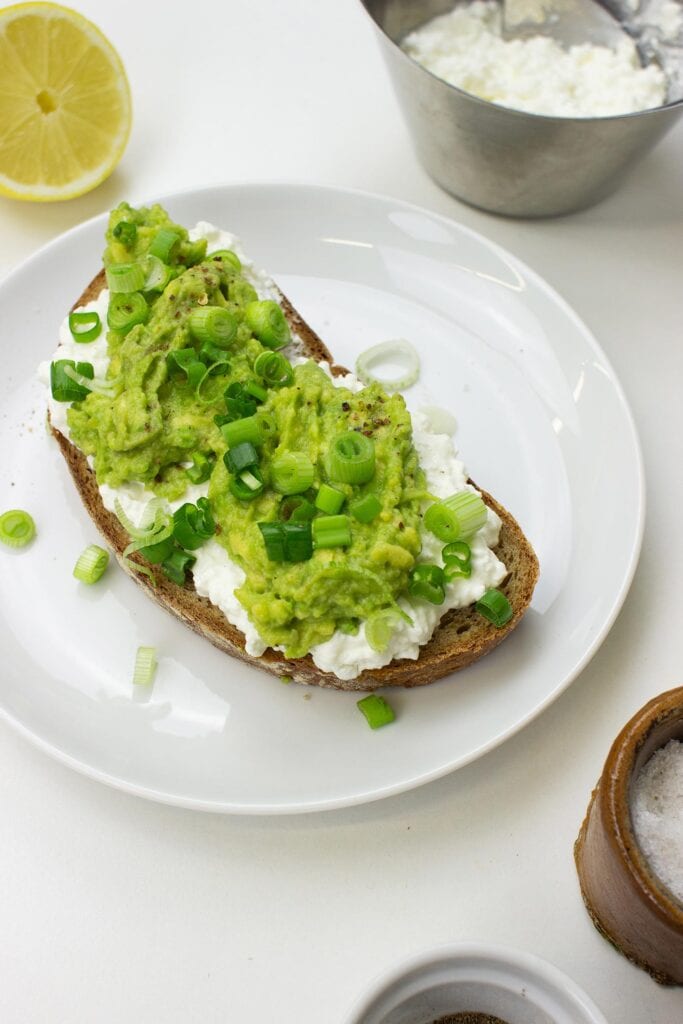 The Avocado Toast with Cottage Cheese is on the plate on a table with salt, pepper, lemon and cottage cheese in the bowl #lemon #green onion | 