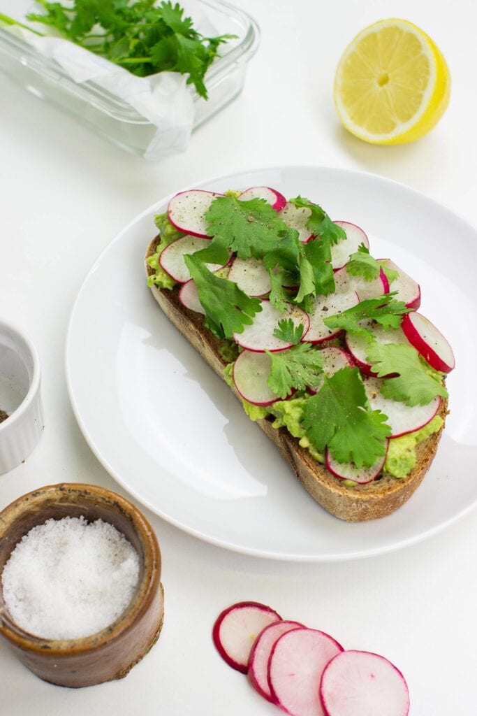 The avocado toast is on the plate on a table with salt, pepper, lemon, chopped radish and cilantro in the lunch box #lemon juice #coriander | 