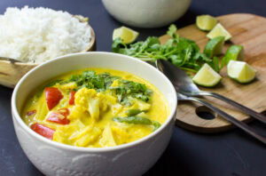A bowl of Vegan Cauliflower Curry is on the dark-blue surface. Next to it there are a wooden bowl with rice and a wooden board with pieces of lime, a bunch of parsley, and two spoons on it | Hurry The Food Up