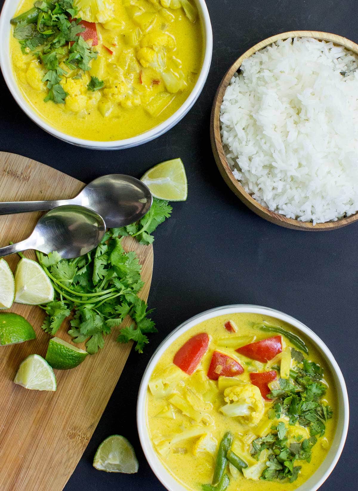 Two bowls of Vegan Cauliflower Curry are on the dark-blue surface. Next to them there are a wooden bowl with rice and a wooden board with pieces of lime, a bunch of parsley, and two spoons on it | Hurry The Food Up