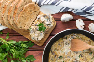 Creamy garlic mushrooms are on the pan. Around it, there are some fresh mushrooms, a bunch of parsley and a wooden board with slices of bread with creamy mushrooms | | Hurry The Food Up