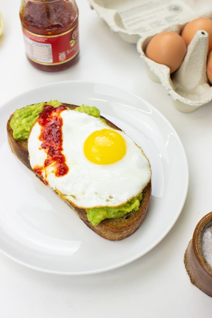 The avocado toast is on the plate on a table with salt, eggs and Sriracha in the jar #bread #avocado | 