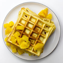 A stack of Orange Buttermilk Waffles, golden and fluffy, topped with fresh orange slices and a dusting of powdered sugar, sits beautifully on a plate.