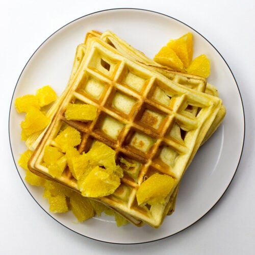 A stack of Orange Buttermilk Waffles, golden and fluffy, topped with fresh orange slices and a dusting of powdered sugar, sits beautifully on a plate.