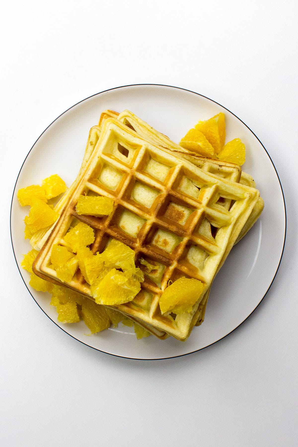 A stack of Orange Buttermilk Waffles, golden and fluffy, topped with fresh orange slices and a dusting of powdered sugar, sits beautifully on a plate.