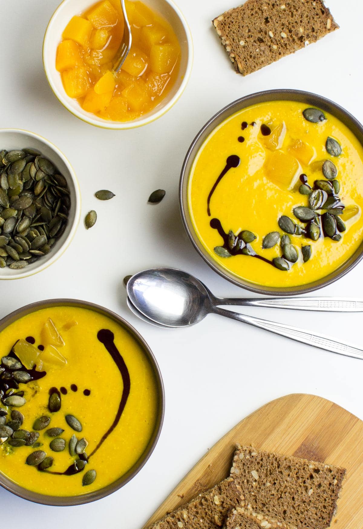 Two bowls with pumpkin and carrot soup. Around them there is a bowl with pumpkin seeds, cut board with bread | Hurry The Food Up