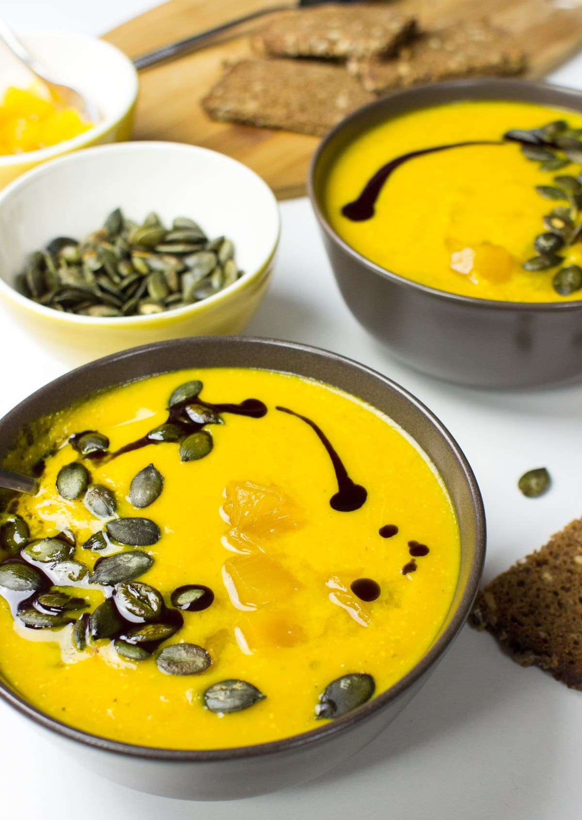 Two bowls with pumpkin and carrot soup. Around them, there is a bowl with pumpkin seeds, a cut board with bread | Hurry The Food Up