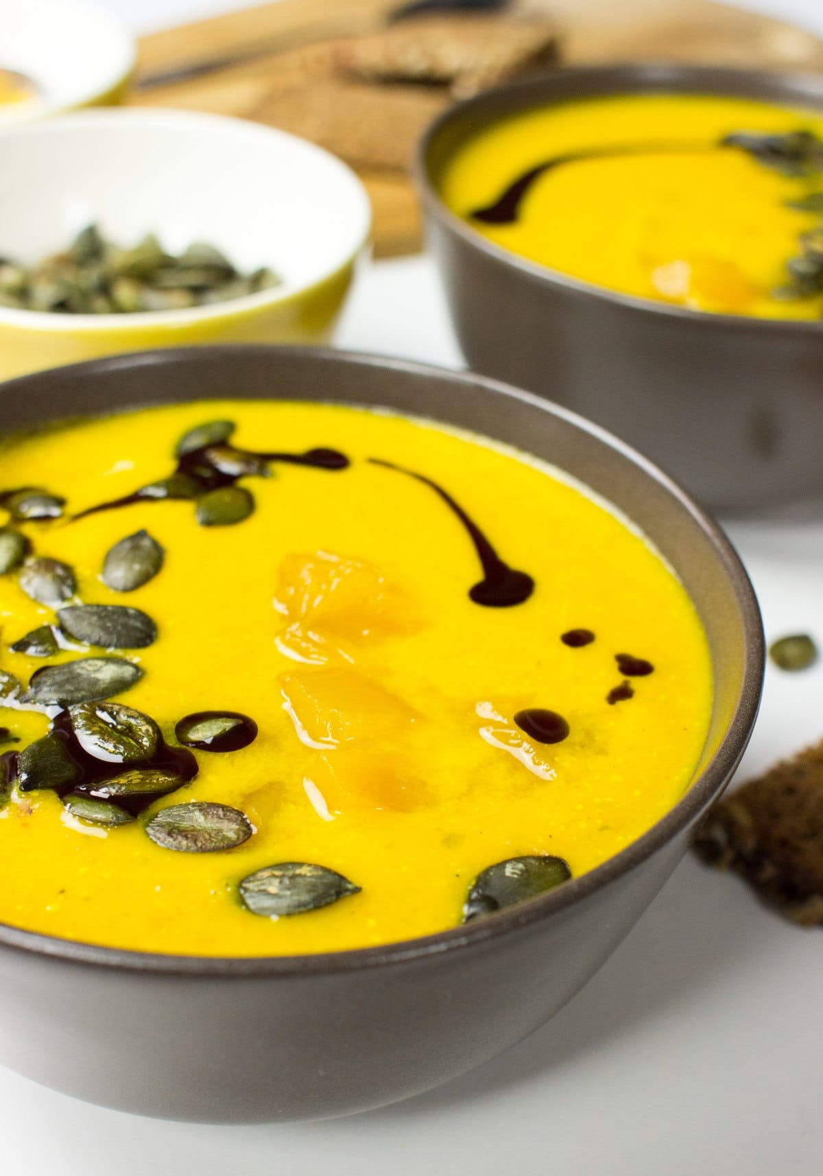 Two bowls with pumpkin and carrot soup. Around them, there is a bowl with pumpkin seeds, a cut board with bread | Hurry The Food Up
