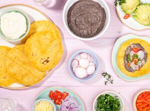 The tortilla is topped with refried beans radish slice, chopped onion, cilantro and grated cheddar cheese that is on the table with all the prepared ingredients | Hurry The Food Up