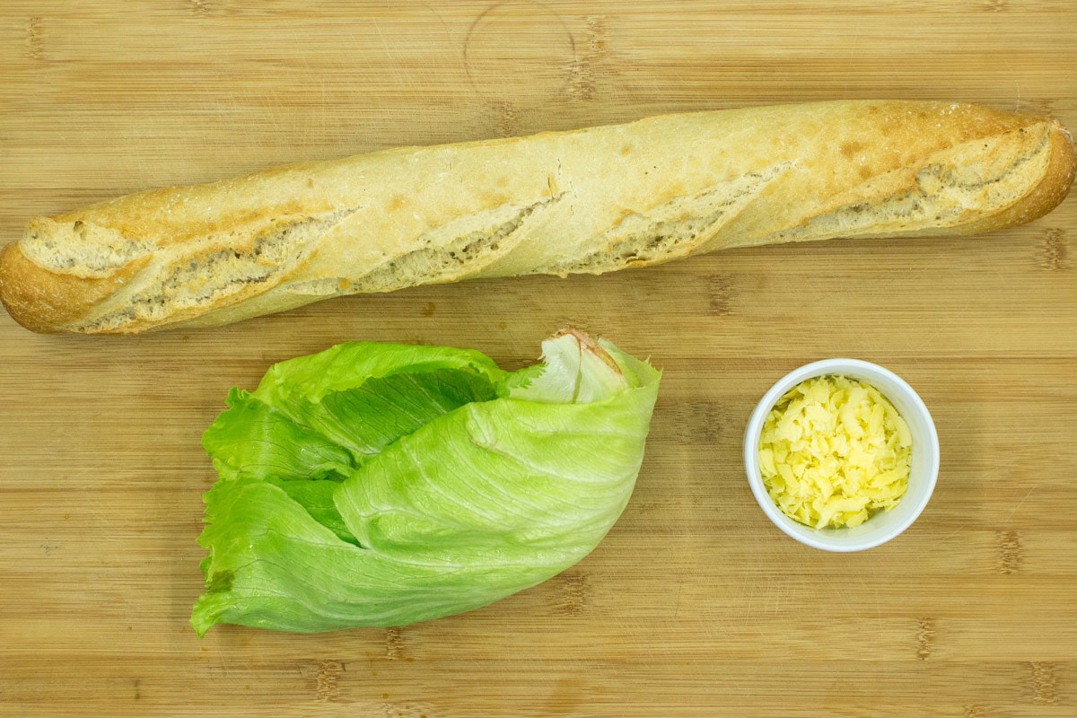Baguette, lettuce, and a white small bowl with cheese are on the wooden surface | Hurry The Food Up