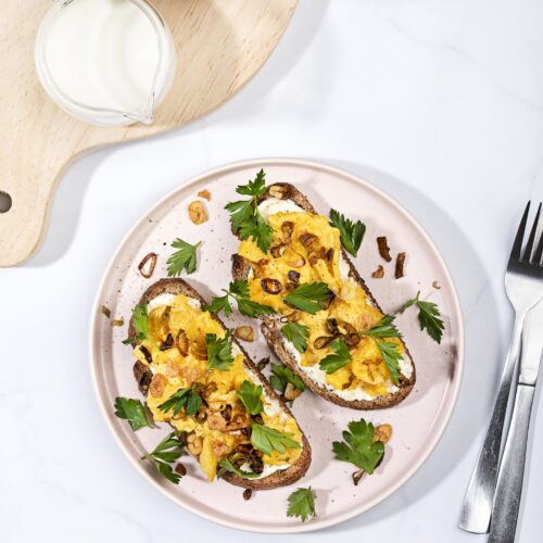 A plate of scrambled eggs on toast from a birds eye view, alongside a cup of coffee and jar of milk, which are place on a chopping board in the left hand corner | Hurry The Food Up