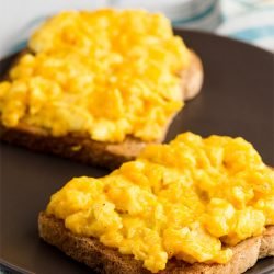 Two pieces of bread with perfect scrambled eggs with cheese lie on the black plate that is on the napkin | Hurry The Food Up
