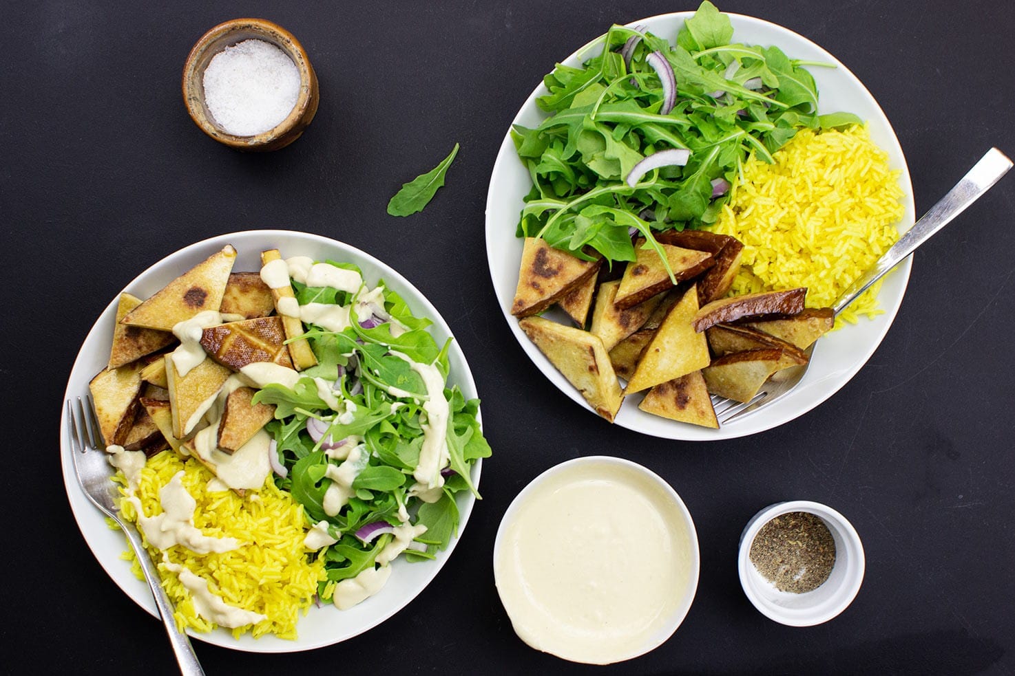 The dish with lamb’s lettuce are on the black table on 2 plates with forks, salt, pepper and hummus #turmeric #basmati rice | 