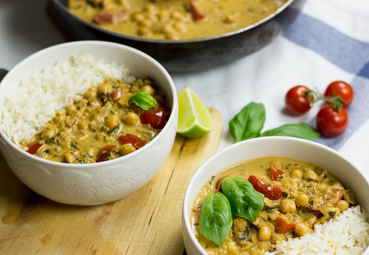 Two bowls with Vegan Chickpea Curry. Next to them, there are some tomatoes, fresh basilic leaves, and a piece of lime | HurryTheFoodUp