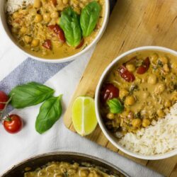 Two bowls with Vegan Chickpea Curry. Next to them, there are some tomatoes, fresh basilic leaves, and a piece of lime | HurryTheFoodUp