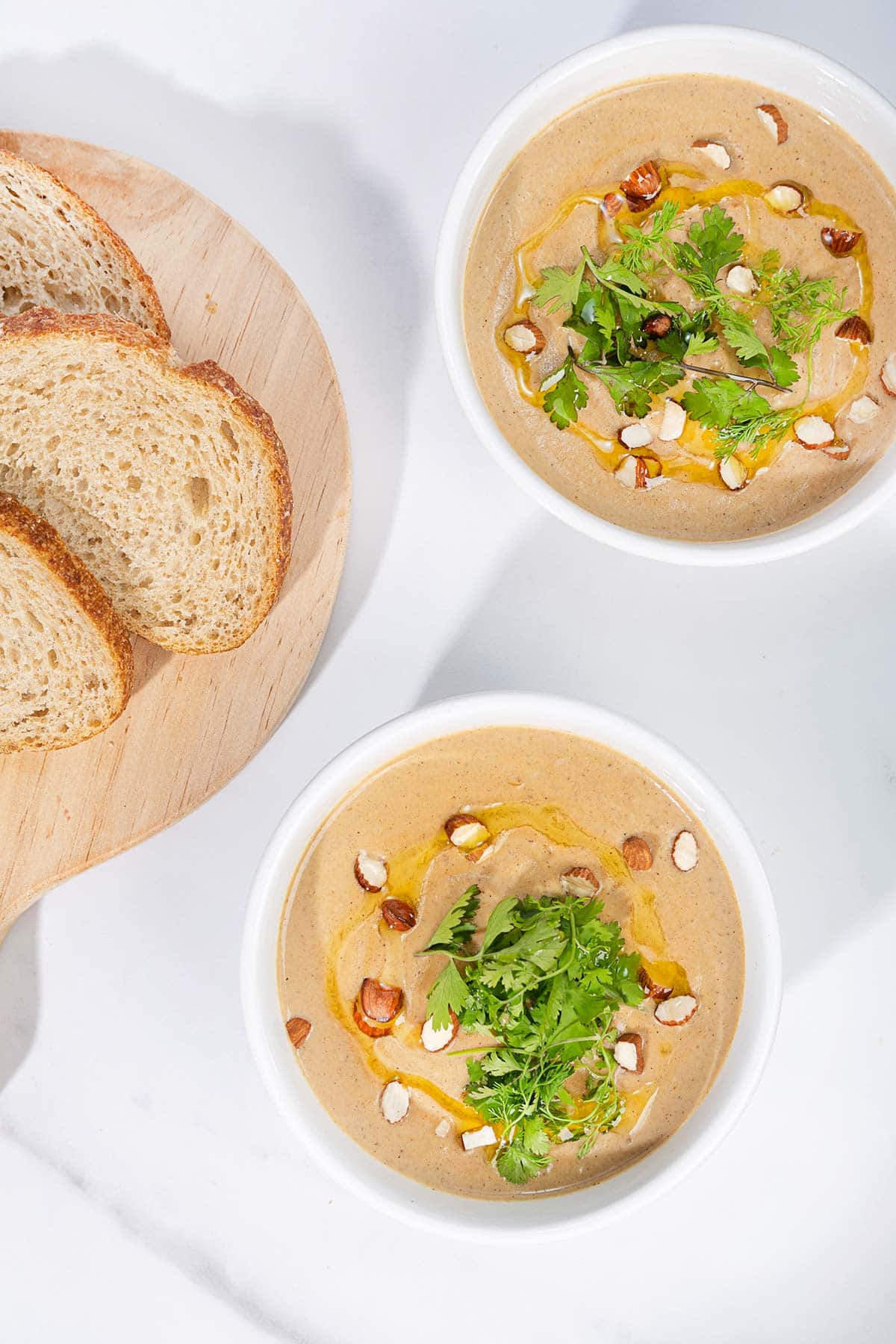 A birds-eye view of a marbled table with two white bowls of creamy vegan cauliflower soup, topped with olive oil, sliced almonds and cilantro. Next to them is a board with some slices of bread | Hurry The Food Up