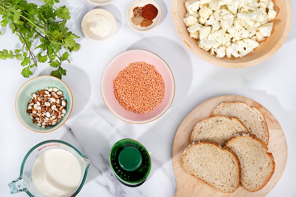 On a marbled table sits a jug of milk, a bowl of sliced almonds, red lentils, and cauliflower florets. Next to them is some cilantro, spices, a bottle of olive oil and a board with some sliced bread on. | Hurry The Food Up