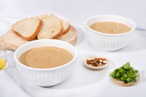 On a marbled table there is a board with slices of bread on it, next to two white bowls of vegan cauliflower soup. There is a small dish of sliced almonds and another with some cilantro. On the left there is a jug of olive oil | Hurry The Food Up