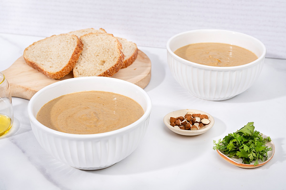 On a marbled table there is a board with slices of bread on it, next to two white bowls of vegan cauliflower soup. There is a small dish of sliced almonds and another with some cilantro. On the left there is a jug of olive oil | Hurry The Food Up
