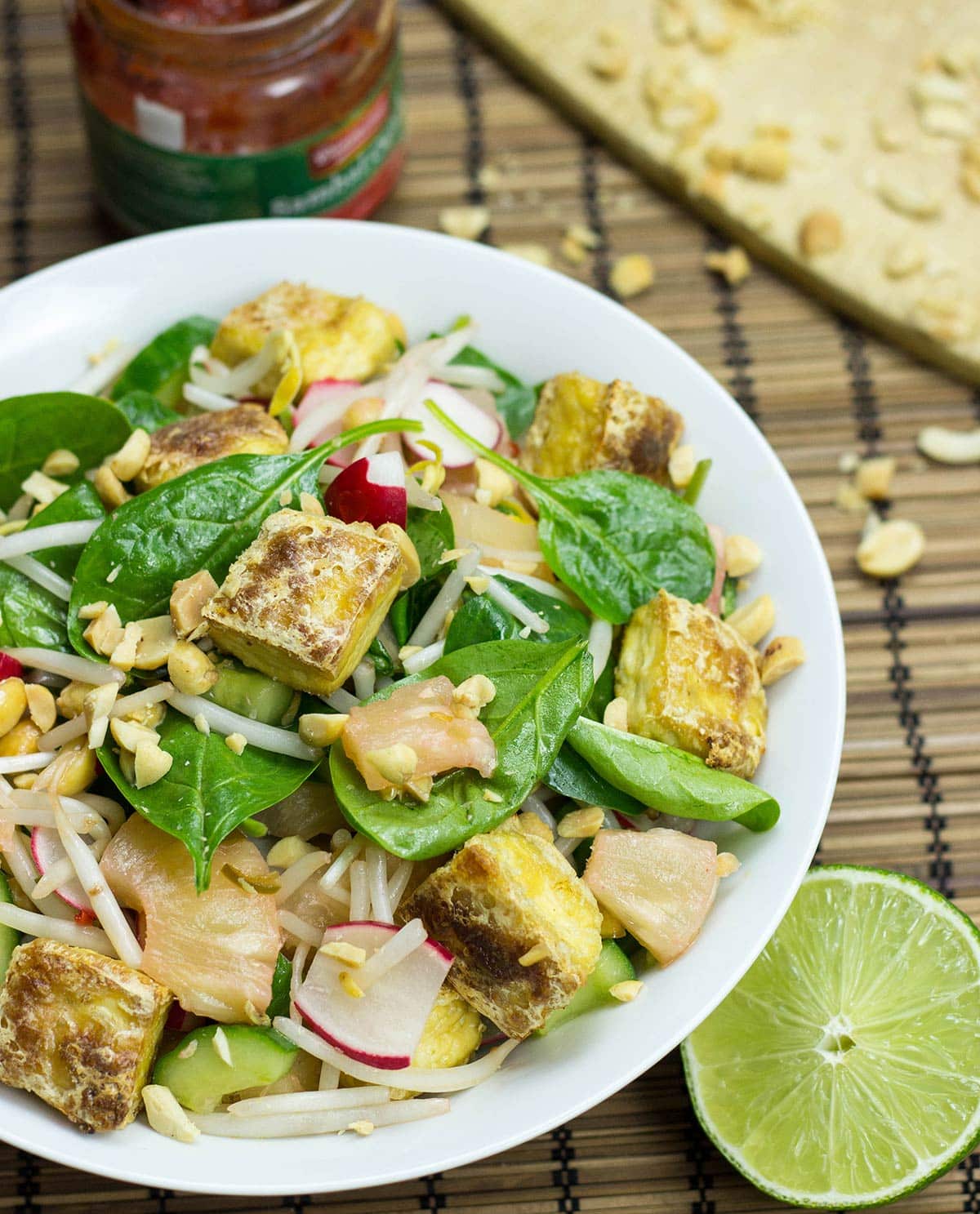 Asian tofu salad is served in a white bowl; a jar of sriracha, some crushed peanuts and a half lime are next to it on the table | Hurry The Food Up