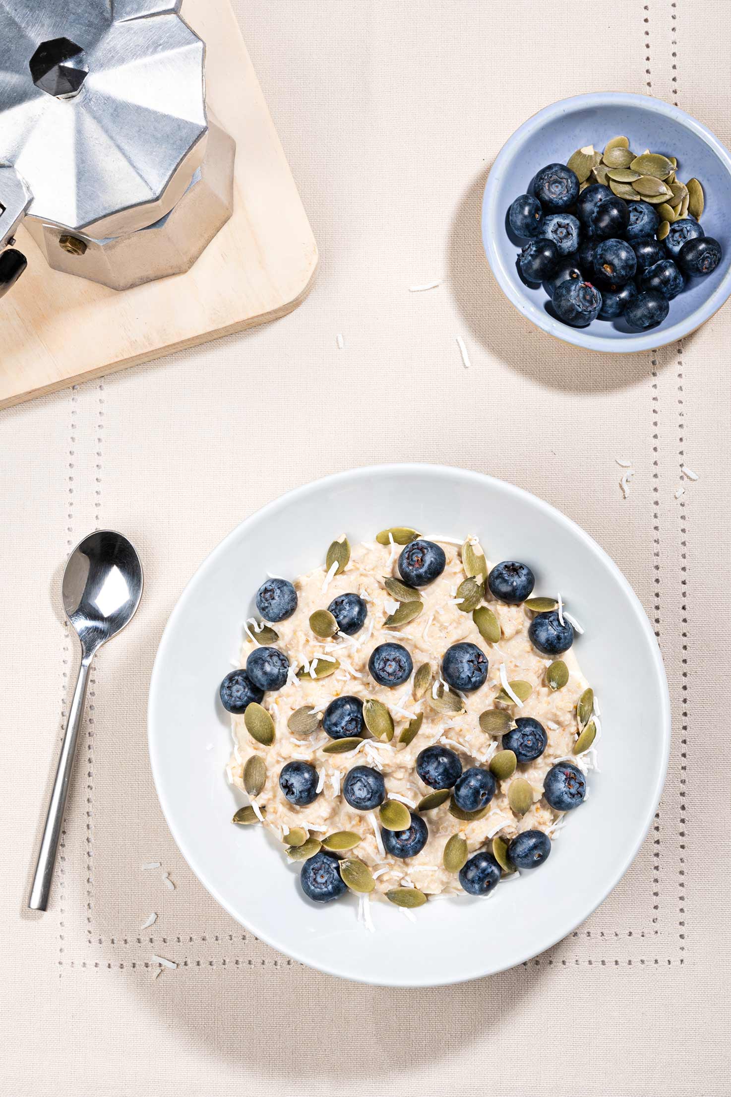 The Bircher muesli is served in a bowl topped with blueberries and pumpkin seeds, there is a spoon on the left | Hurry The Food Up