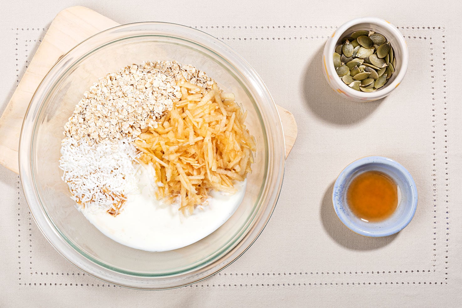 Oats, grated apple, coconut flakes and yougurt are in a bowl not mixed up, pumpkin seeds and maple syrup are in separate small bowls on the right | Hurry The Food Up