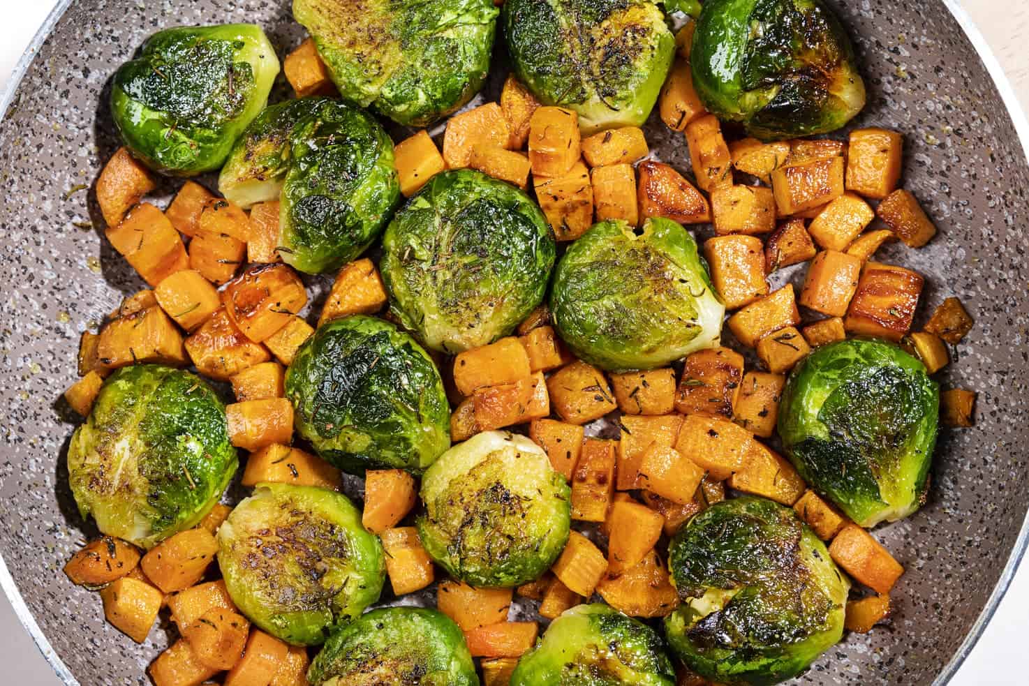 Smashed brussel sprouts and cubed carrots are in a frying pan with herbs. They are browned and becoming caramelised.