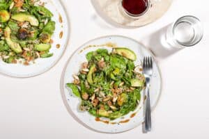 Two plates of smashed brussel sprouts salad with quinoa are on a white surface, with a cup of water & a jug of dressing. | Hurry The food Up