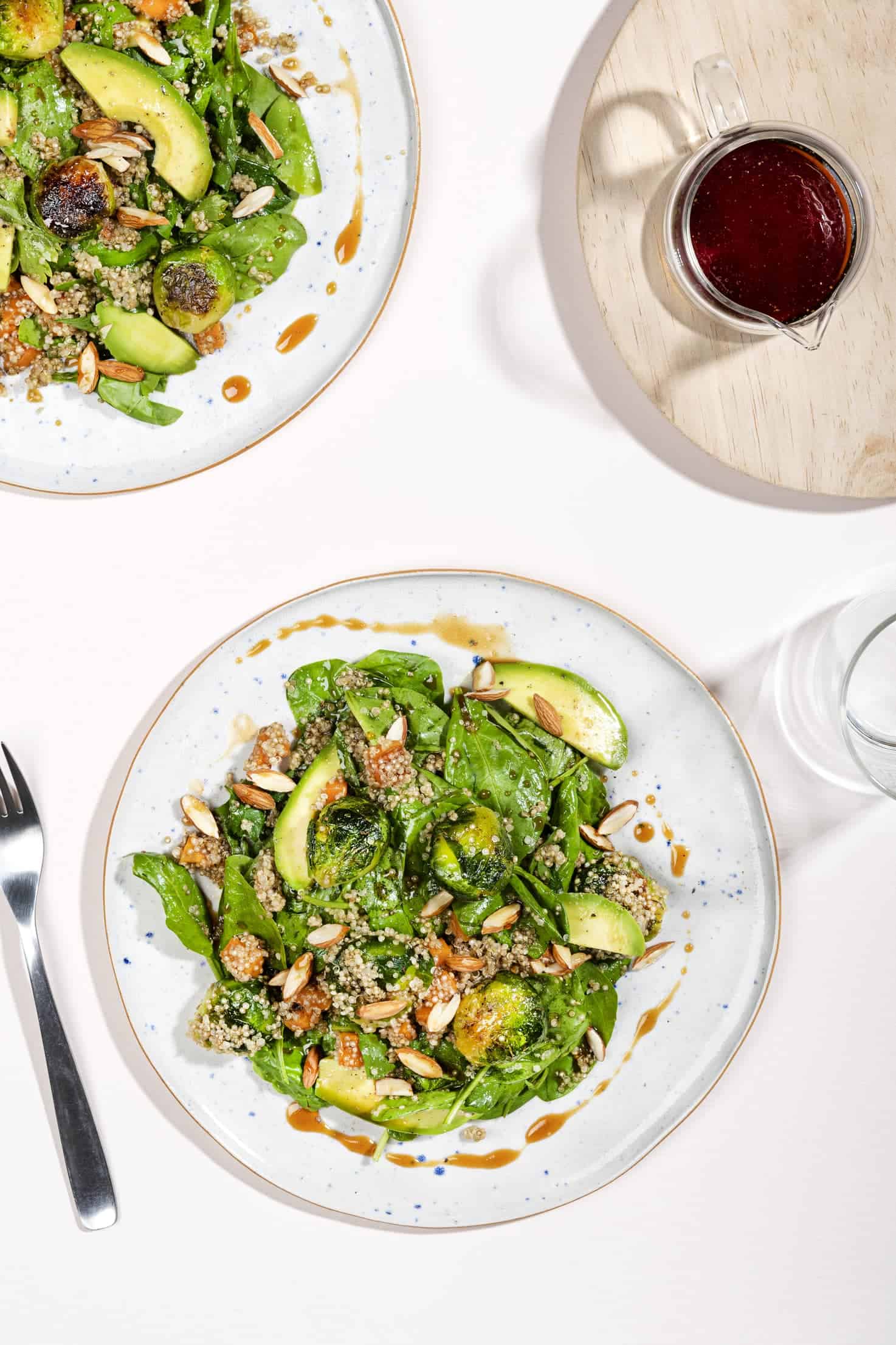 Two plates of brussel sprouts salad with quinoa from birds eye view. Nearby on a wooden board is a jug of dressing.