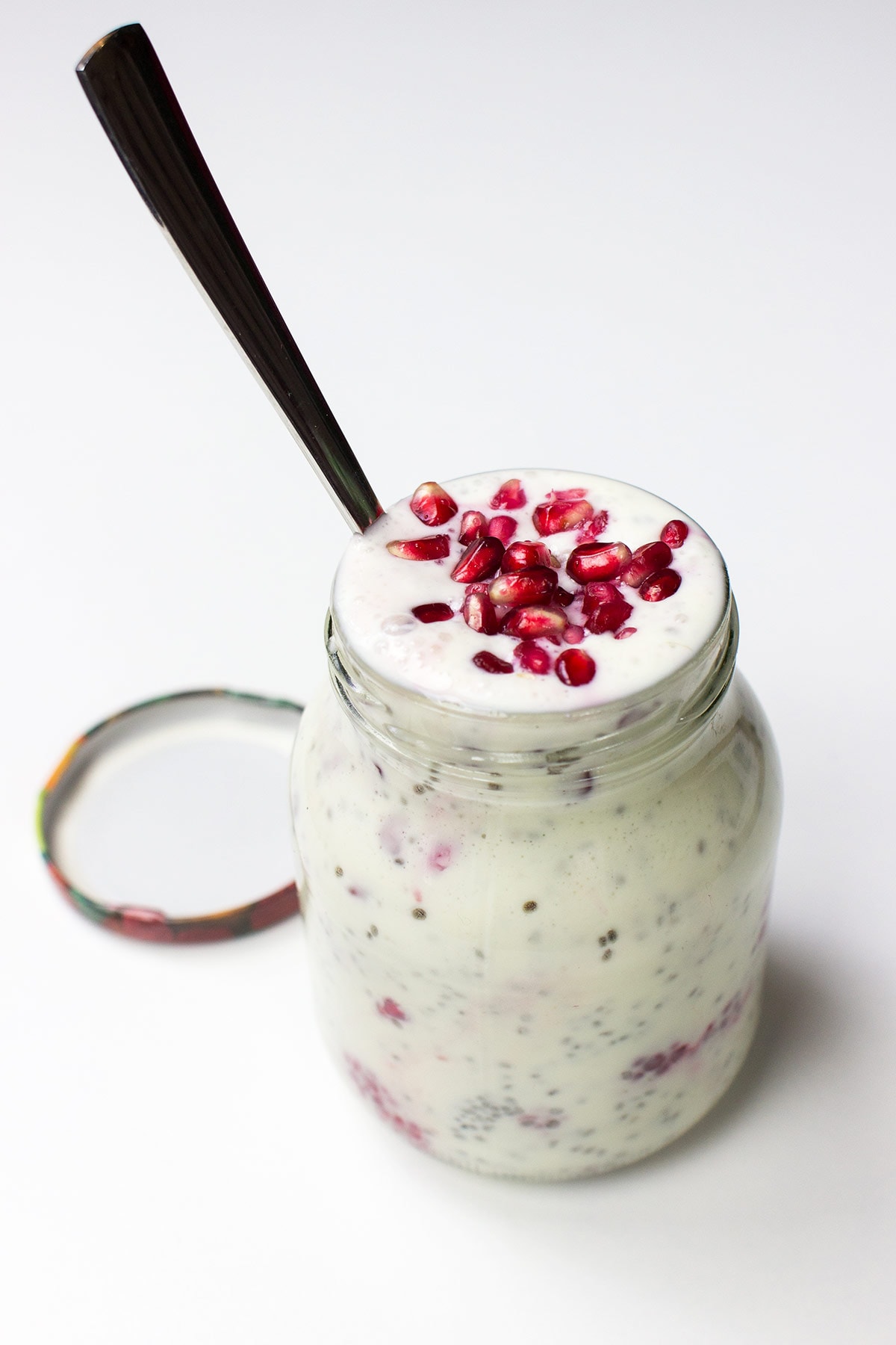 A glass jar filled with a Chia Yoghurt Bowl topped with pomegranate seeds and raspberries, with a spoon inserted and the lid placed beside it on a white surface.