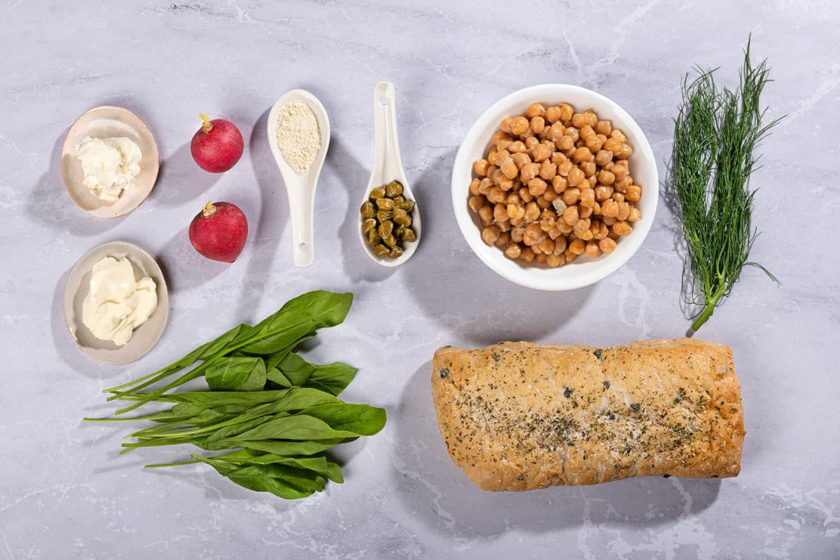 Chickpea salad sandwich ingredients like chickpeas, sour cream, low fat cream cheese, capers, baby spinach, radishes, bread etc. are on a marble surface | Hurry The Food Up