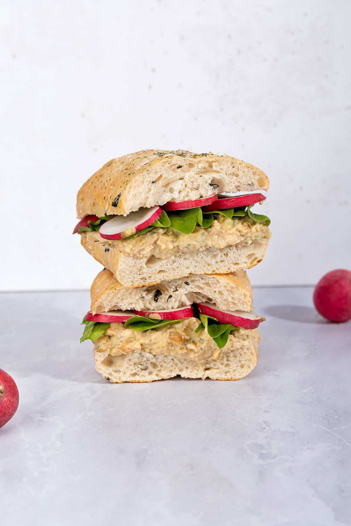 Two chickpea salad sandwiches stacked on top of each other on a marble surface with two radishes next to them | Hurry The Food Up