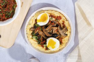 A cream bowl of polenta with mushrooms sundried-tomatoes and basil with eggs on top sits on a marbled surface between a wooden chopping board and a cream cloth| Hurry The Food Up