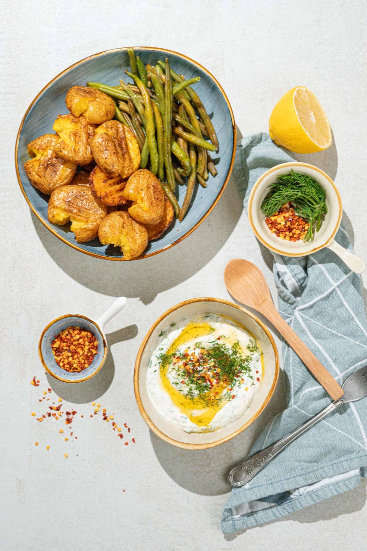 Bird's-eye view of smashed potatoes and green beans which is on the grey surface. Near it, there are smaller bowls with feta dipping sauce, sweet paprika flakes, dill etc. | Hurry The Food Up