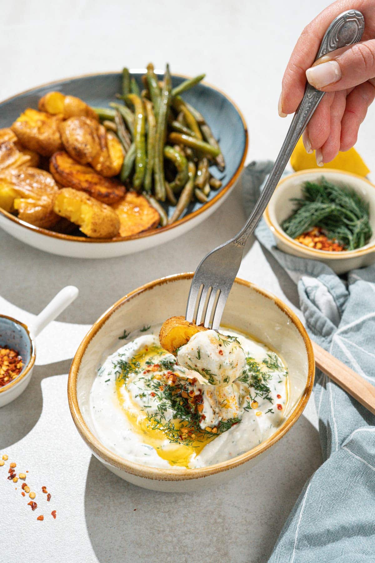 A bowl with smashed potatoes and green beans is on the grey surface. Near it, there are smaller bowls with feta dipping sauce, sweet paprika flakes, dill etc. | Hurry The Food Up