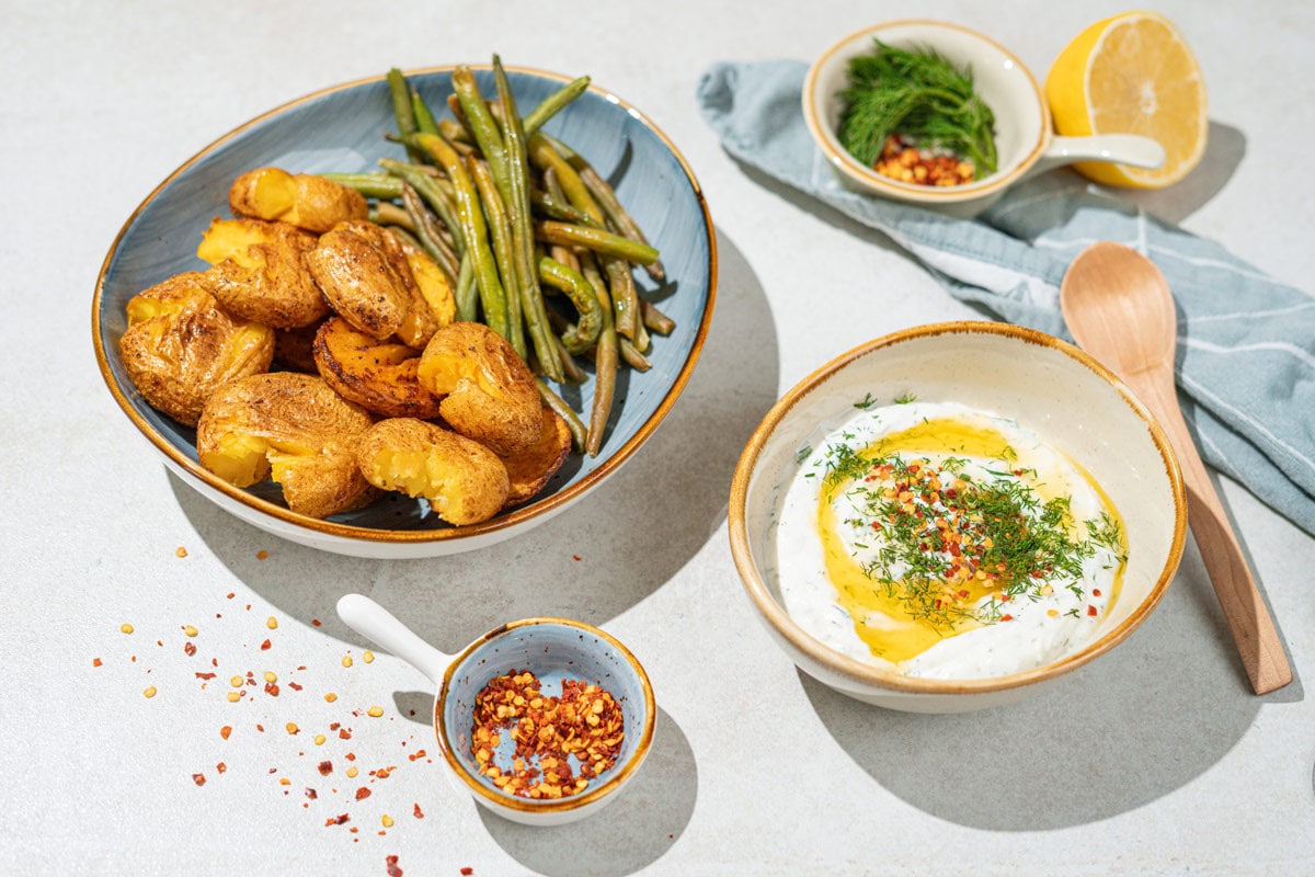 A bowl with smashed potatoes and green beans is on the grey surface. Near it, there are smaller bowls with feta dipping sauce, sweet paprika flakes, dill etc. | Hurry The Food Up