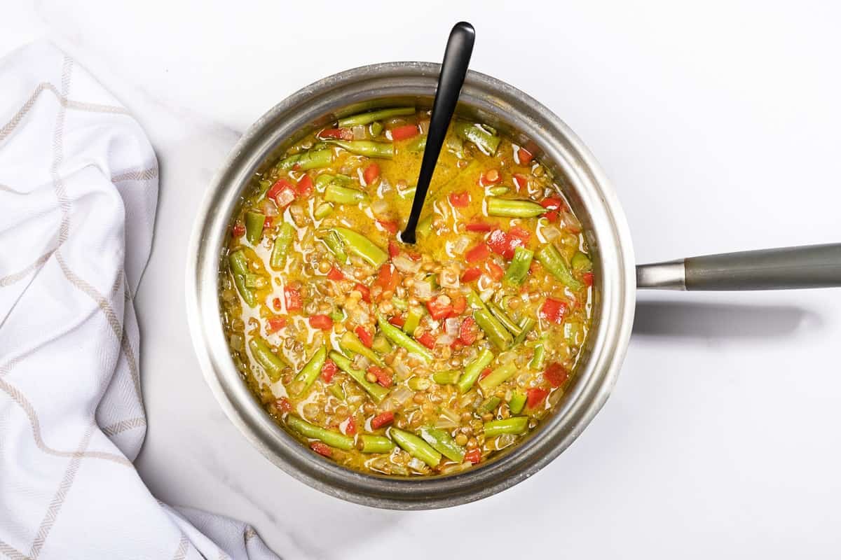 A saucepan full of curried lentil soup with coconut being cooked, with a big stirring spoon in it. | Hurry The Food Up