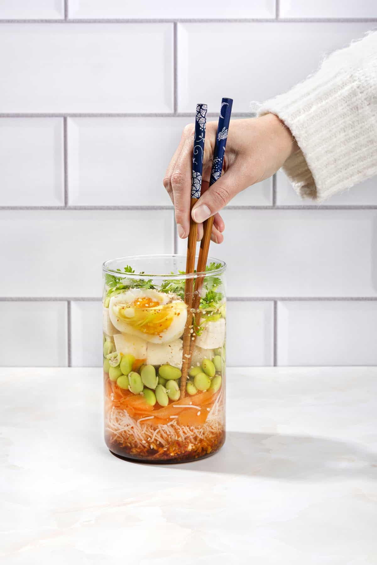 A person dips a pair of chopsticks into a jar of noodles, vegetables, tofu and egg in a stock based soup | Hurry The Food Up