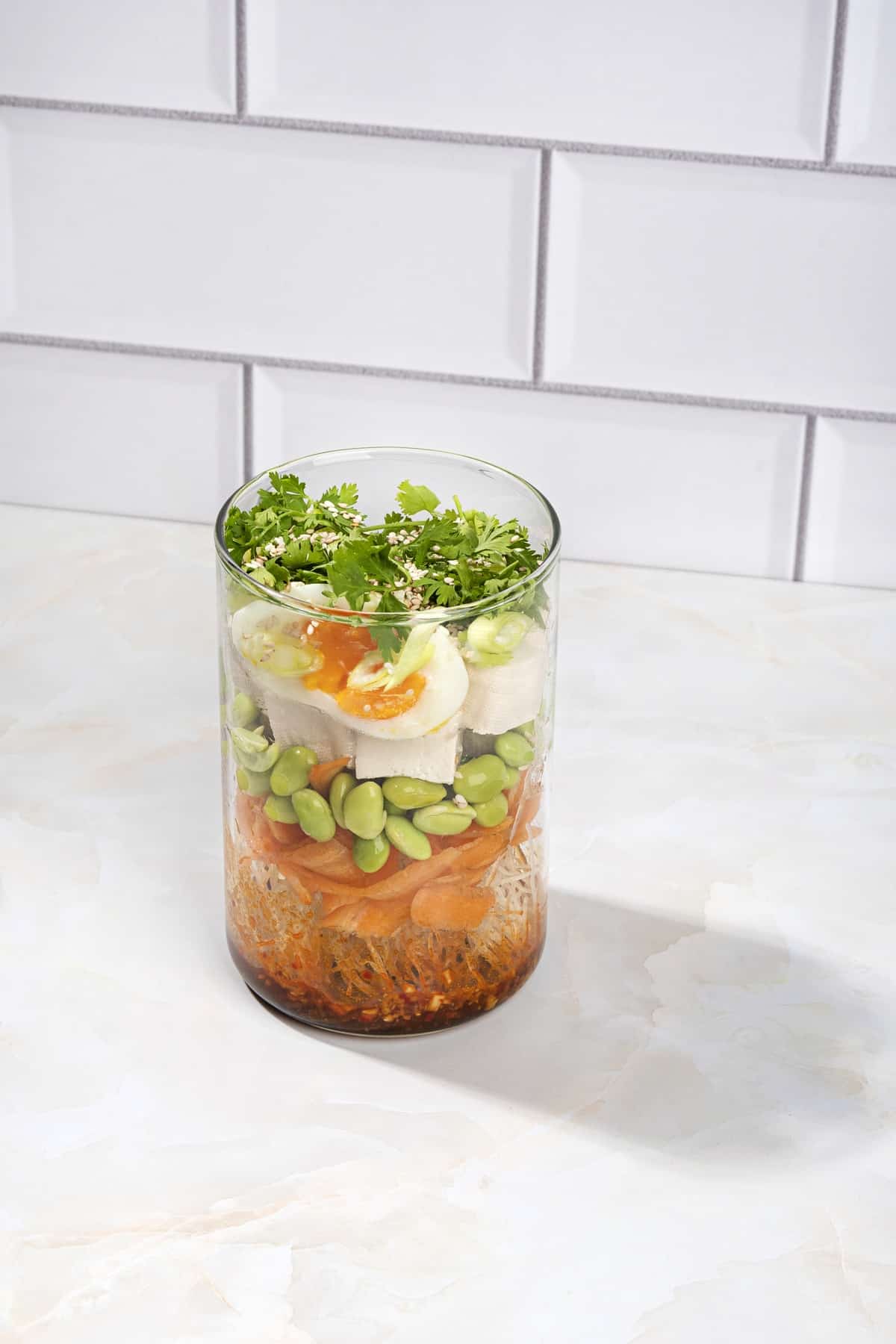 A jar layered with seasoning noodles, veggies, tofu and eggs is open on a white kitchen counter | Hurry The Food Up