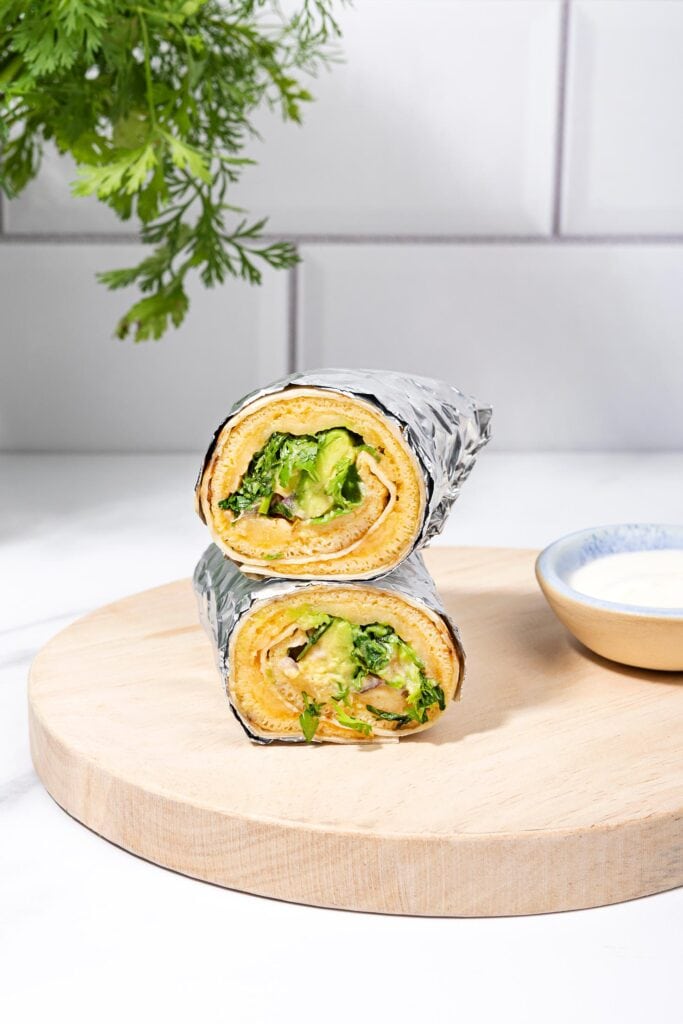 Two breakfast egg wraps in foil on a cutting board, a small bowl of sour cream near them | Hurry The Food Up