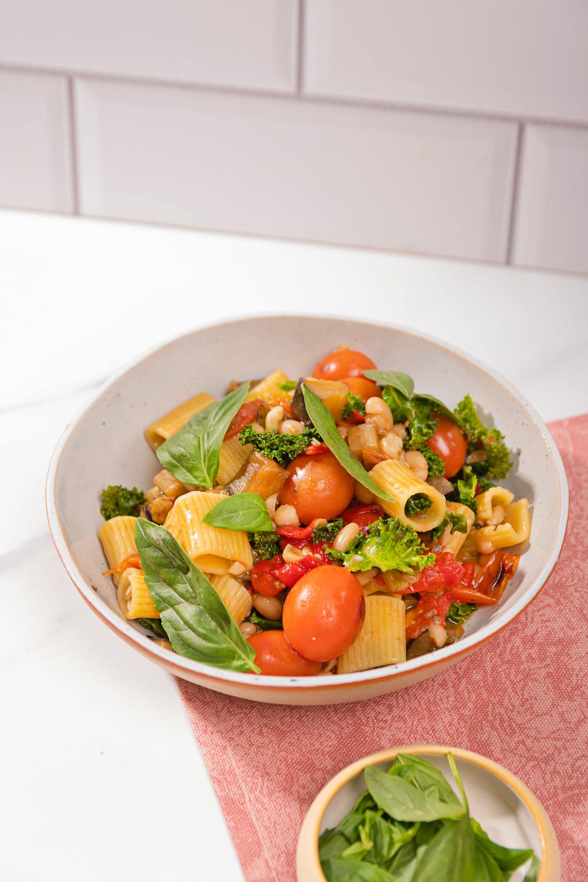 Saucy eggplant tomato pasta is in the white plate on the pink napkin that is on the table. Next to it there is a plate with basil | Hurry The Food Up