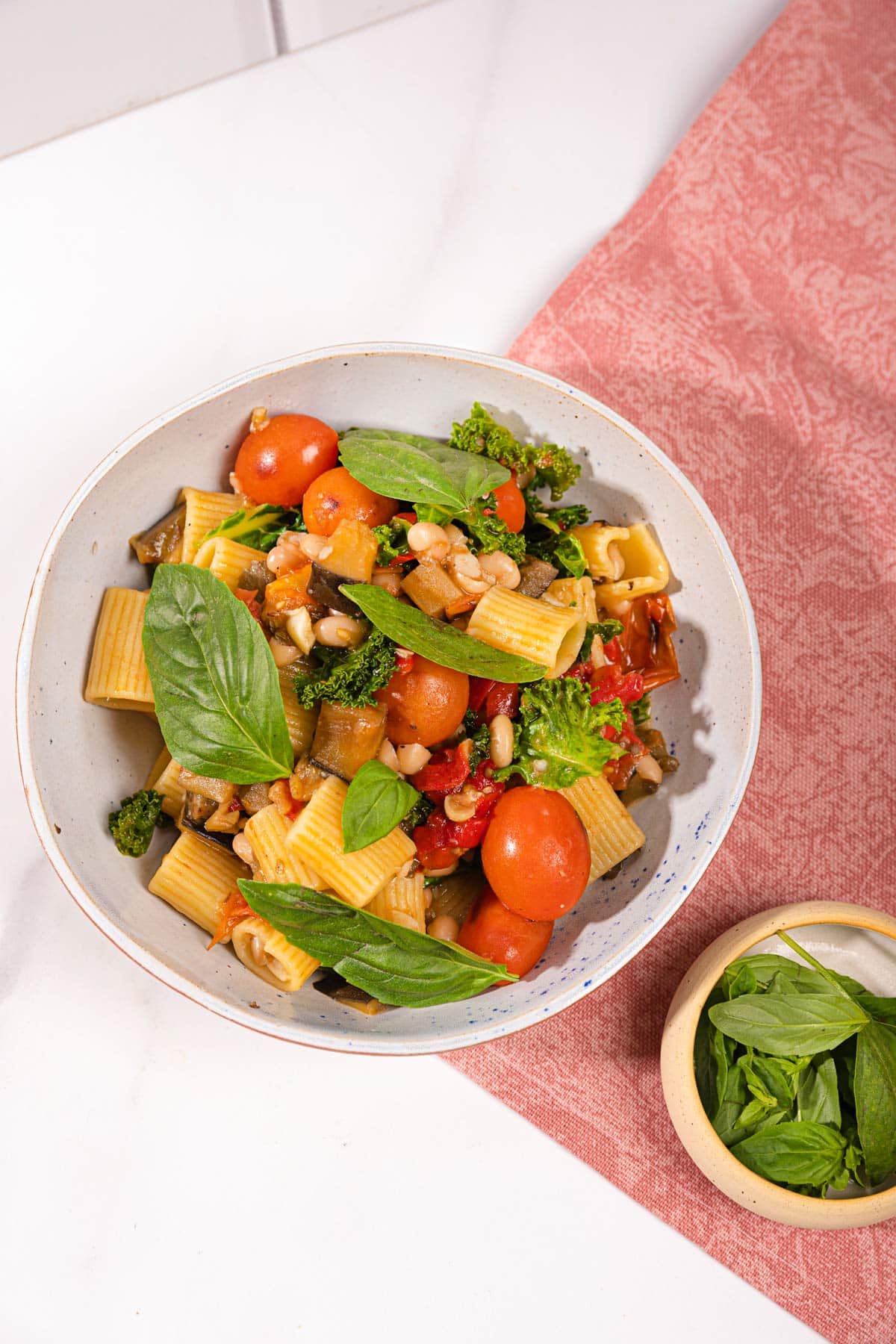 Saucy eggplant tomato pasta is in the white plate on the pink napkin on the table. Next to it there is a plate with basil | Hurry The Food Up