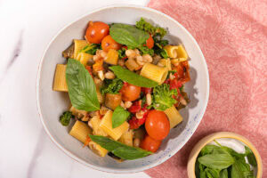 Bird's-eye view of saucy eggplant tomato pasta is in the white plate on the pink napkin. Next to it there is a plate with basil | Hurry The Food Up