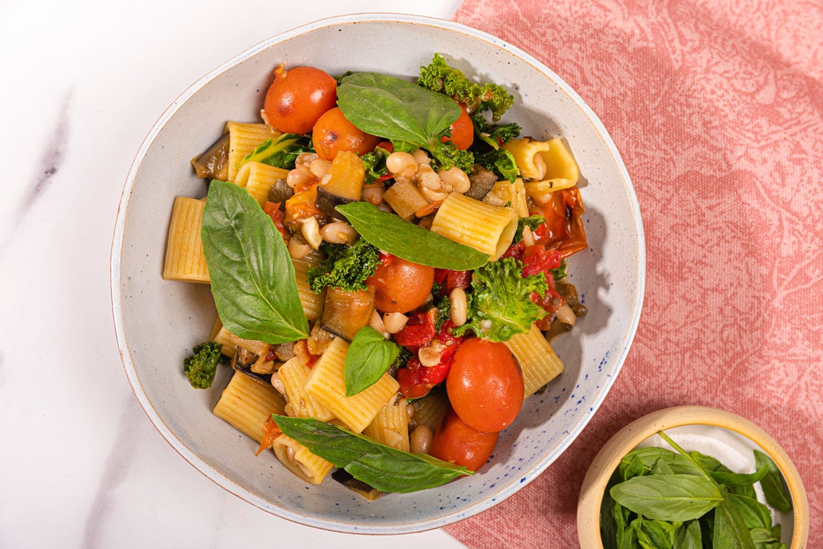 Bird's-eye view of saucy eggplant tomato pasta is in the white plate on the pink napkin. Next to it there is a plate with basil | Hurry The Food Up