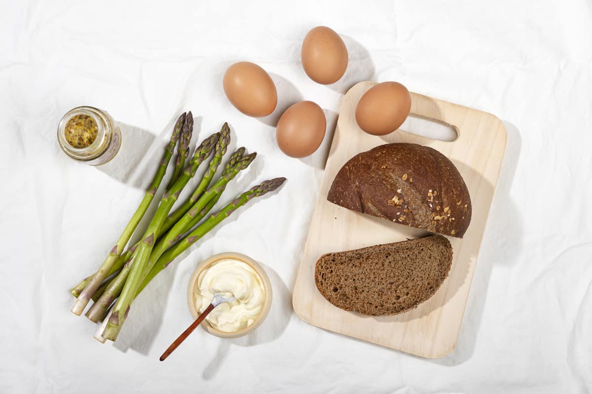 A bunch of asparagus, 4 boiled eggs, a jar of dijon mustard and a loaf of dark bread on a white surface | Hurry The Food Up