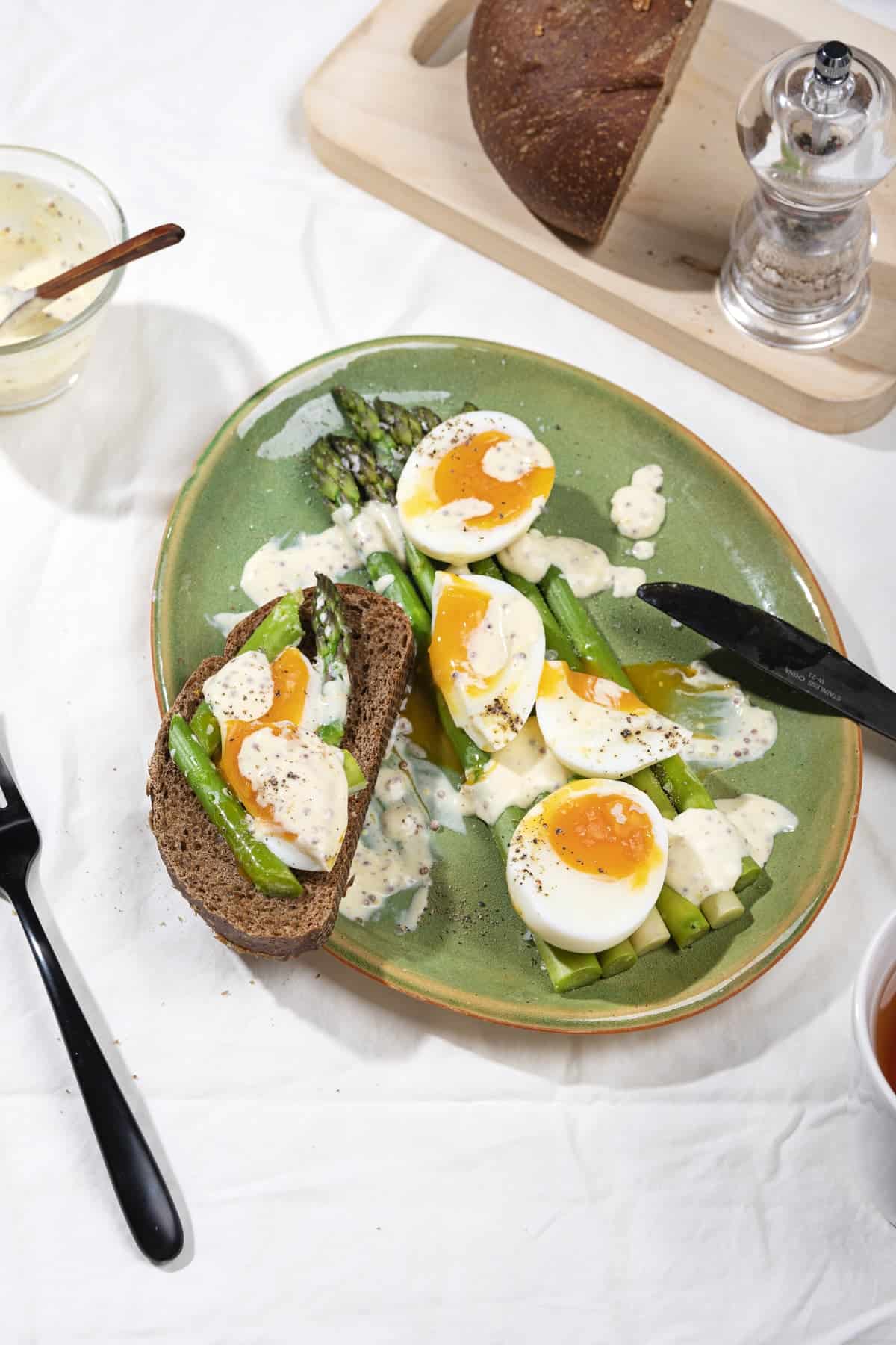 A plate of asparagus, jammy eggs, sour cream sauce and dark bread in the process of being eaten for breakfast | Hurry The Food Up