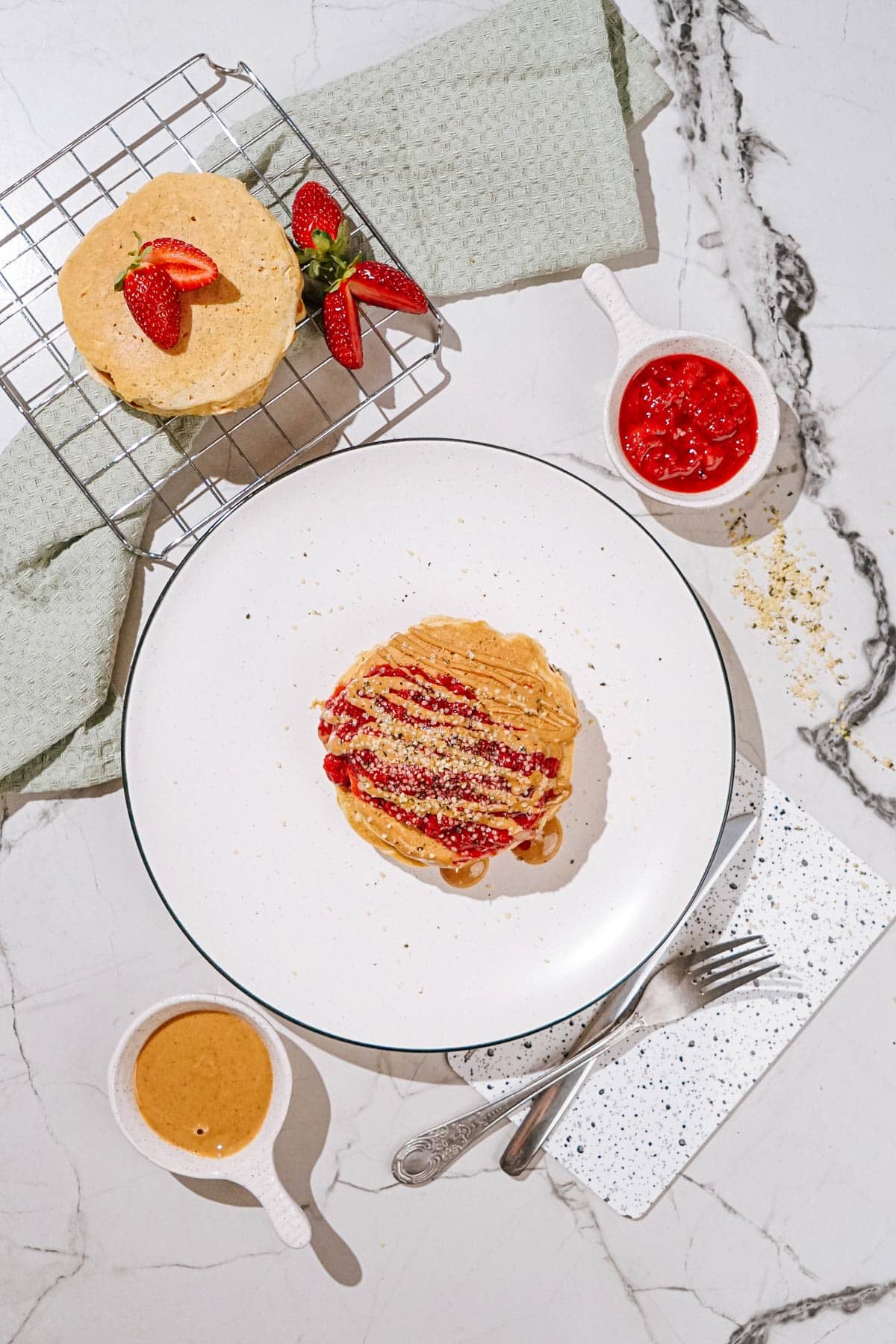 Bird's-eye view of the plate with fluffy protein powder pancakes drizzled with nut butter and sprinkled with hemp seeds is on a light surface. Near it, there are some small dishes with strawberry compote, nut butter, silverware etc | Hurry The Food Up
