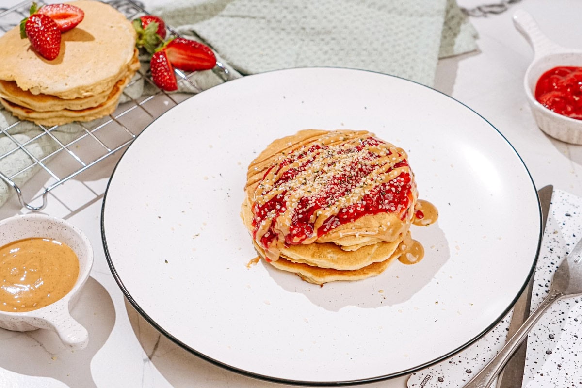 The plate with fluffy protein powder pancakes drizzled with nut butter and sprinkled with hemp seeds is on a light surface. Near it, there are some small dishes with strawberry compote, nut butter, silverware etc | Hurry The Food Up