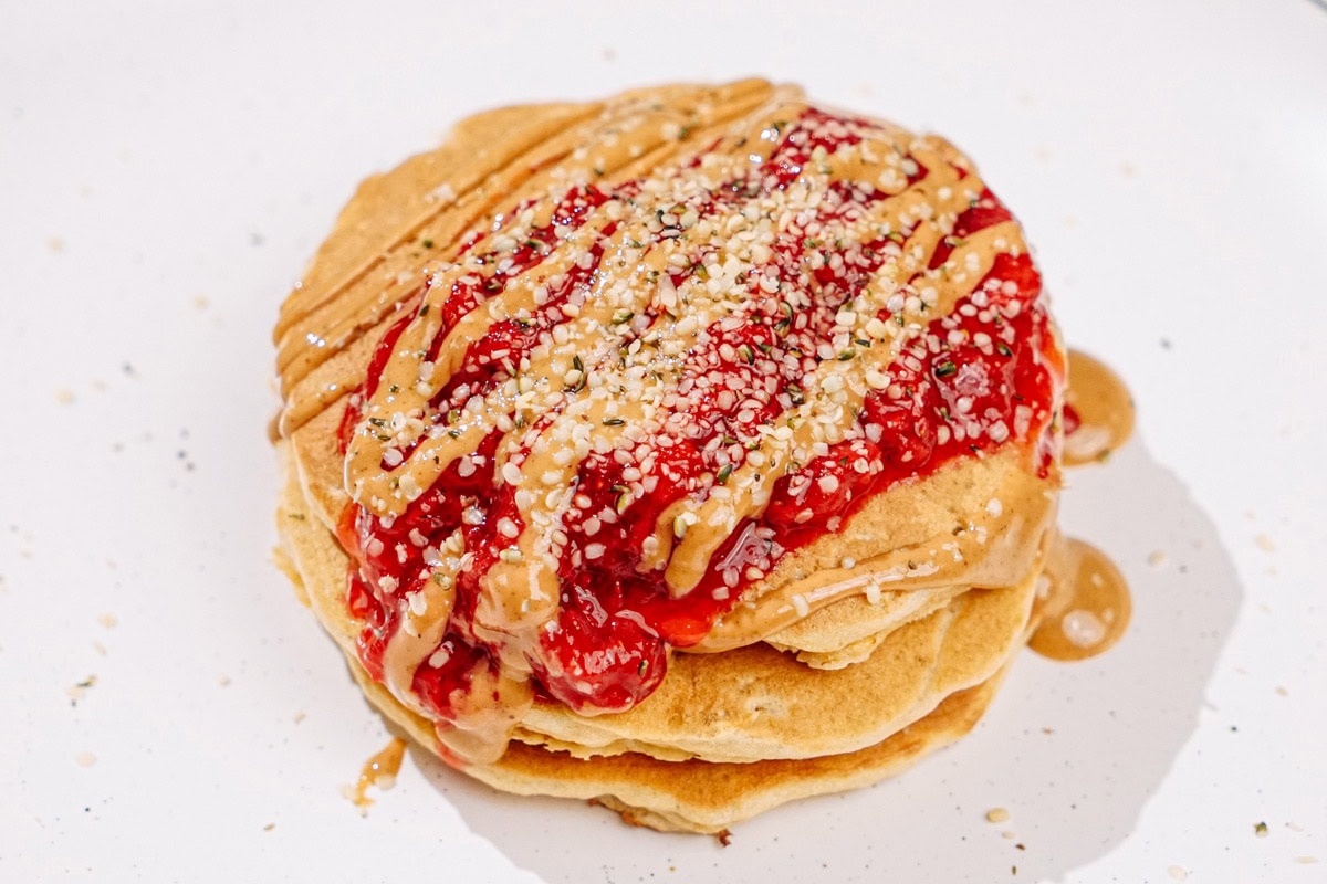 The process of pouring strawberry compote over stacked pancakes, drizzling some nut butter, and sprinkling with hemp seeds | Hurry The Food Up