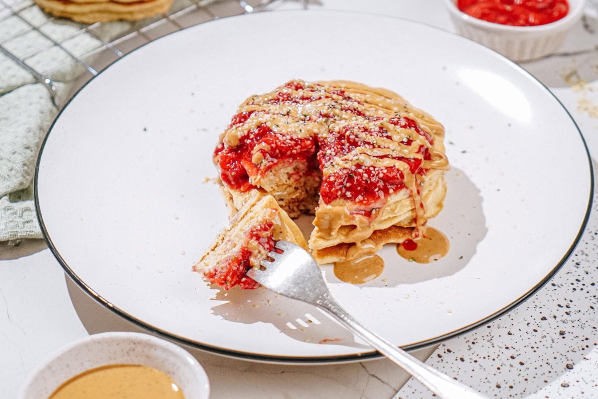The plate with fluffy protein powder pancakes, which are drizzling nut butter, and sprinkling hemp seeds is on the light surface | Hurry The Food Up